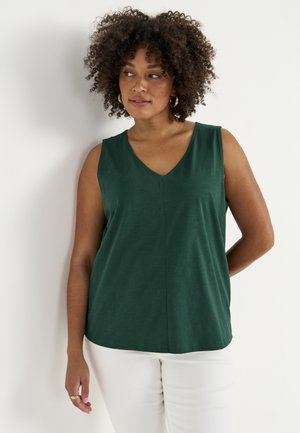 Kvinde med krøllet hår iført guldringe øreringe, en mørkegrøn ærmeløs top og hvide bukser, stående foran en hvid baggrund.