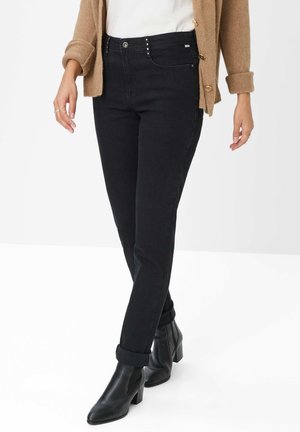Schwarze Jeans im Slim-Fit-Design, Fünf-Taschen-Stil und dekorativen Metallnieten am Bund, kombiniert mit schwarzen Stiefeletten.