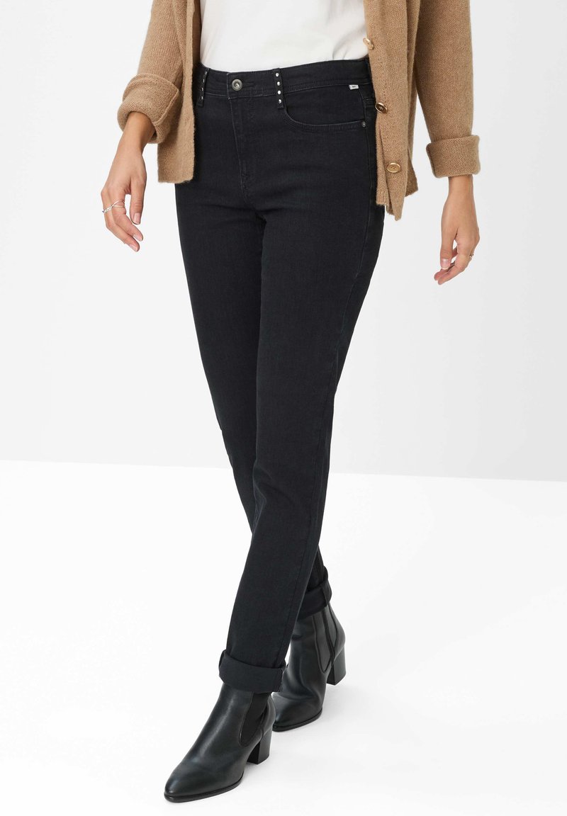 Jeans in denim nero con un design aderente, stile a cinque tasche e borchie decorative in metallo alla vita, abbinati a stivali neri alla caviglia.