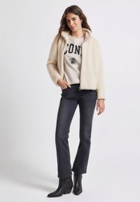 Kurtka z kremowego futra ekologicznego, biała bluza z czarnymi literami, ciemne dżinsy i czarne botki na obcasie. Modelka stoi z rękami w kieszeniach kurtki.