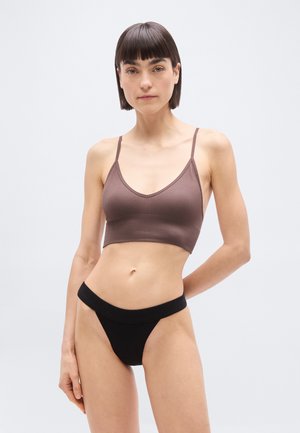 Γυναίκα με κοντά σκούρα μαλλιά που φοράει καφέ κοντό τοπ και μαύρο εσώρουχο με ψηλή μέση, στέκεται μπροστά σε απλό ανοιχτό φόντο.