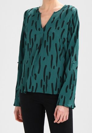 Blouse verte à manches longues avec des rayures abstraites noires, col en V et coupe ample portée avec un pantalon noir.