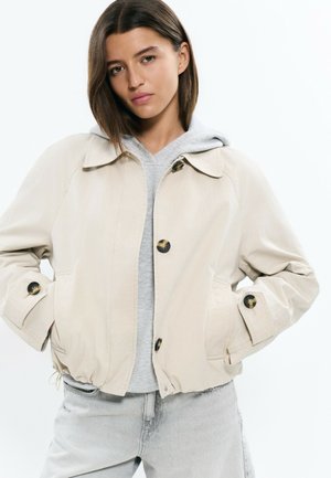 Mujer con cabello castaño liso que lleva una chaqueta beige abotonada sobre una sudadera con capucha gris y jeans gris claro, con las manos en los bolsillos de la chaqueta, expresión neutral.