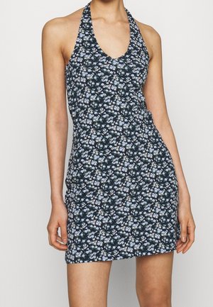 Mini sukienka z kołnierzem halter, czarne tło i mały wzór kwiatowy w jasnoniebieskim i białym kolorze, noszona przez stojącą osobę.