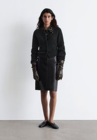 Schwarze flauschige Strickjacke, schwarzer Lederrock im Krokodilmuster, braune Lederhandschuhe und schwarze Slipper, kombiniert mit einem gemusterten Hemd mit Kragen.