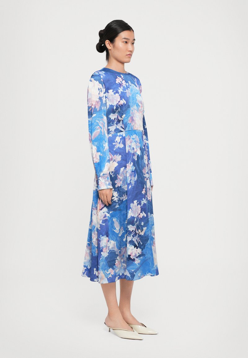 Vrouw met donker haar in een lage bun, gekleed in een langemouwen, blauw bloemdress tot op de kuit en crèmekleurige puntige schoenen, staande tegen een effen achtergrond.