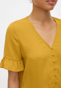 Blusa gialla senape con maniche corte a volant, scollo a V e piccoli dettagli di bottoni sul davanti. Tessuto liscio e leggero.