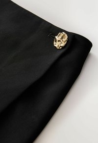Tessuto nero con una superficie liscia, caratterizzato da un bottone decorativo dorato con un design intricato, posato pianamente su uno sfondo chiaro.