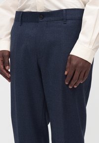 Homme portant un pantalon de costume texturé bleu foncé et une chemise crème à manches longues, les mains reposant sur les côtés, montré de la taille au milieu de la cuisse.
