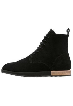 Bottine noire en daim à lacets avec une semelle en caoutchouc plate et un talon empilé en bois, avec une languette à l'arrière.