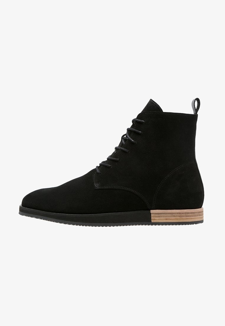 Bottine noire en daim à lacets avec une semelle en caoutchouc plate et un talon empilé en bois, avec une languette à l'arrière.