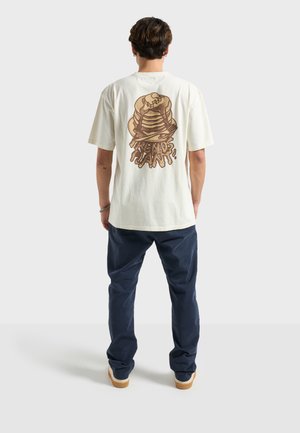 Kremfarvet bomulds-T-shirt med en stor tegneseriestil-illustration af stablede pandekager på bagsiden, med detaljer af dryppende sirup.