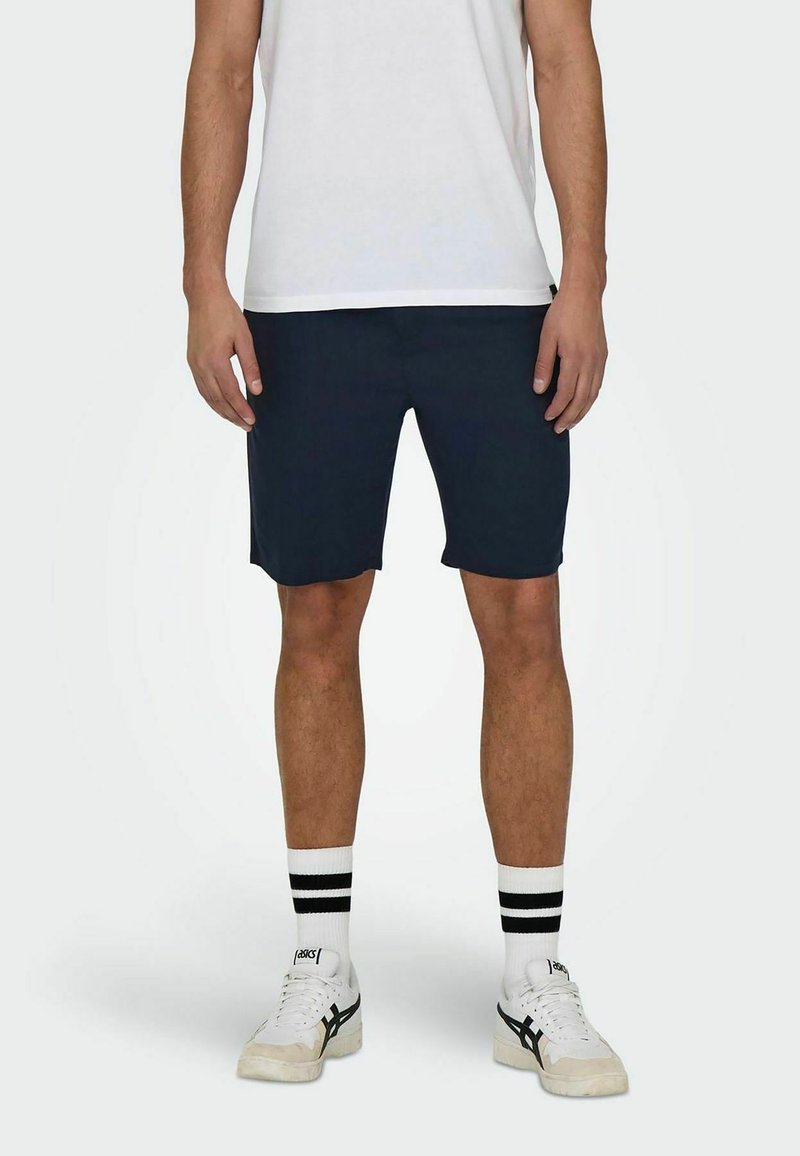 Pantaloni corti blu con taglio dritto, realizzati in tessuto leggero, abbinati a una t-shirt bianca, calze a righe e sneaker casual.