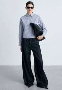 Striped button-up shirt in light blue and white, paired with wide-legged dark denim trousers. Model carries a black leather clutch bag.