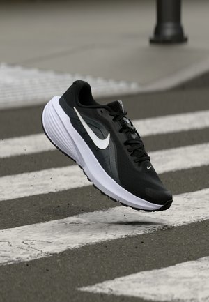 Zapatilla de running Nike en blanco y negro flotando sobre un paso de peatones en una carretera asfaltada con fondo desenfocado.