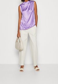 Top sin mangas de satén púrpura, pantalones anchos blancos, bolso de mano en beis claro y sandalias de beis con tiras trenzadas.