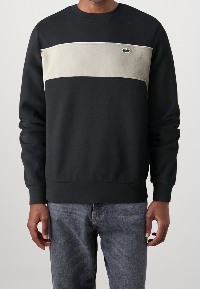 Homme portant un sweat-shirt noir avec un panneau beige sur la poitrine et un petit logo crocodile vert, assorti à un jean bleu, debout devant un fond uni.