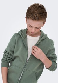 Niño con sudadera verde y camiseta blanca mirando hacia abajo mientras cierra parcialmente la sudadera en un fondo claro.