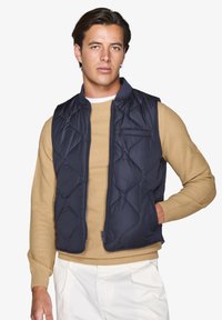 Marineblau gesteppte Weste mit Stehkragen, Reißverschluss und seitlichen Taschen, kombiniert mit einem beige strukturierten Strickpullover.