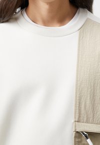 Sudadera de algodón blanco con panel lateral texturizado en beige. Presenta un cuello redondo acanalado y detalles de bolsillo con cremallera.