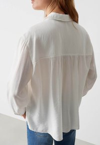 Camicia bianca a maniche lunghe realizzata in un tessuto morbido e leggero. Presenta un colletto a punta e dettagli arricciati sul retro, abbinata a jeans blu.