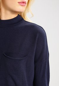 Mörkblå stickad tröja med hög krage, förslagen med en liten bröstficka, texturerad tyg och en lös passform.