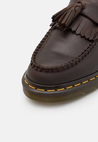 Mocassin en cuir marron foncé avec glands, couture moc toe et semelle épaisse en caoutchouc jaune sur fond blanc.