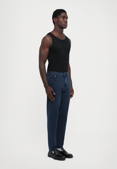 Man standing wearing a black sleeveless top, dark blue jeans, white socks, and black leather shoes on a plain light background.