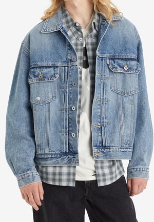 Personne portant une veste en jean bleue sur une chemise à carreaux grise et un T-shirt blanc, accompagnée d’un pantalon noir, présentée des épaules aux hanches.