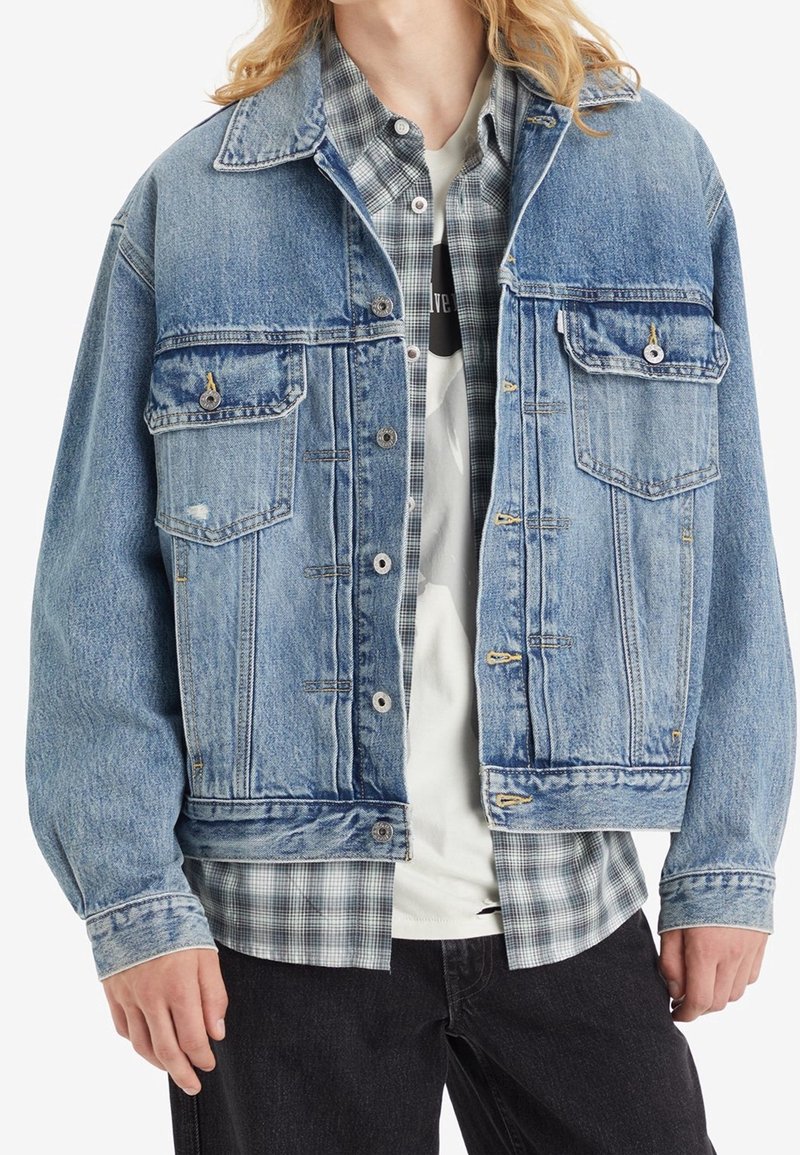 Personne portant une veste en jean bleue sur une chemise à carreaux grise et un T-shirt blanc, accompagnée d’un pantalon noir, présentée des épaules aux hanches.