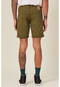 Shorts khaki con orlo arrotolato e texture in denim, dotati di tasche posteriori e di una patch in cuoio in vita, abbinati a scarpe nere e calzini verdi.