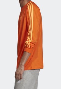 Camisa naranja de manga larga con tres rayas amarillas en las mangas, con un cuello redondo y un corte relajado. Fabricada en un tejido ligero.