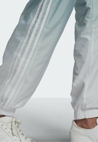 Ljusblå träningsbyxor med vit tre-randig design. Har elastiska muddar och en slät, glansig yta. Kombinerade med vita sneakers.