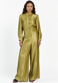 Vrouw die staat in een glanzend olijfgroen shirt met lange mouwen en knopen, gecombineerd met een wijde broek en zwarte open teen hakken, tegen een witte achtergrond.