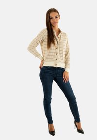 Cardigan de color beige claro con rayas horizontales, cierre de botones y puños acanalados, combinado con jeans skiny de mezclilla oscura y tacones negros de punta.