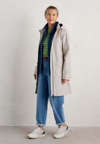 Veste imperméable gris clair avec capuche, zip intégral à l'avant et poignets ajustables, portée par-dessus une veste en denim bleu et un top rayé vert.