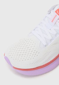 Baskets blanches avec tige en mesh, accents violets et rouges, bout arrondi, semelle texturée et lacets plats. Logo de la marque dans un coin.