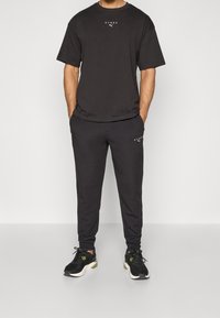Camiseta negra de manga corta con logo, combinada con pantalones jogger negros a juego. Presenta una cinturilla elástica y tobillos ajustados. Lleva puestas unas deportivas negras.