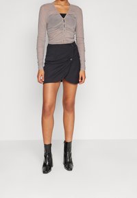 Langärmliges, gerafftes graues Oberteil mit Knöpfen, kombiniert mit einem schwarzen Wickelrock mit einem Knopfdetail und zwei Taschen, getragen mit schwarzen High Heels.