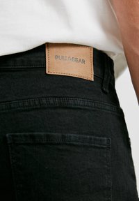 Gros plan de la partie arrière de la taille d’un jean noir avec une étiquette en cuir beige cousue portant l’inscription "PULL&BEAR".