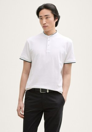 Polo bianco con colletto rialzato, dotato di tre bottoni e bordi neri lungo le maniche. Indossato con pantaloni scuri e una cintura.