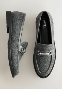 Grijze platte loafers met zwarte synthetische trim en een zilverkleurig decoratief hardwareaccent. Dikke zwarte zool, ronde neus en klassiek ontwerp.