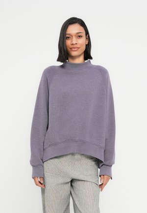 Paarse sweatshirt met een hoge hals, raglanmouwen en een cropped zoom. Het materiaal lijkt zacht met een gestructureerde afwerking. Gecombineerd met lichte, patroonbroeken.