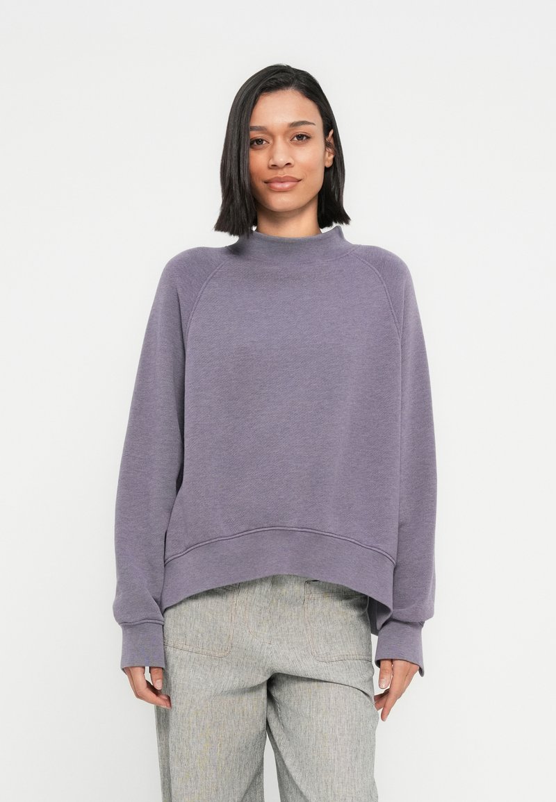 Purple sweatshirt with a high neckline, raglan sleeves, and a cropped hem. Material appears soft with a textured finish. Paired with light patterned pants.