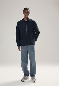 Námornícka bunda na zips s dvoma náprsními vreckami, v kombinácii s ľahko modrými voľnými džínsami a sivými teniskami. Hladká textúra, ležérny dizajn.