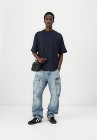 Tricou oversized navy, blugi cargo de culoare albastru deschis cu multiple buzunare, geantă crossbody neagră și adidași negri cu dungi albe.