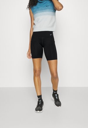 Jonge vrouw die een blauw-wit mouwloos topje draagt, zwarte Nike fietsshorts, zwarte Nike sokken en zwarte hardloopschoenen, staand.