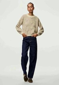 Kremfarget strikket genser med diamantmønster, rund hals og senkede skuldre, kombinert med mørkeblå jeans med rette ben og brune sko.