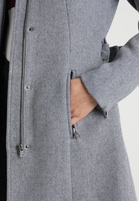 Cappotto grigio in misto lana con chiusura zip, tasche laterali e superficie strutturata. Dettaglio di una mano appoggiata vicino a una tasca.