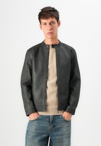 Jeune homme aux cheveux courts et foncés portant une veste en cuir noire sur un pull beige et un jean bleu, les mains dans les poches, expression neutre, fond blanc.